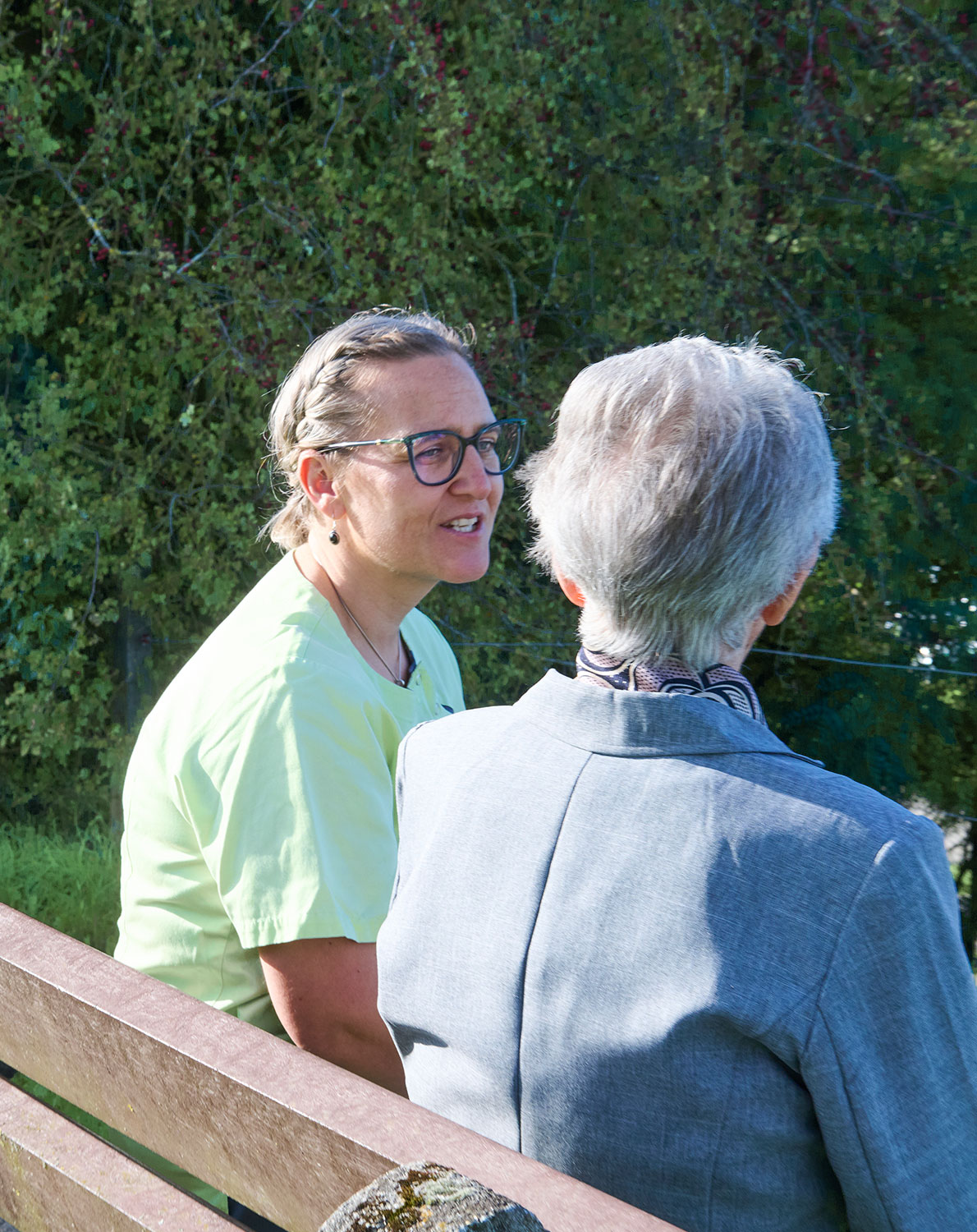 Eine Pflegerin und eine Bewohnerin sitzen auf einer Gartenbank im Freien und unterhalten sich