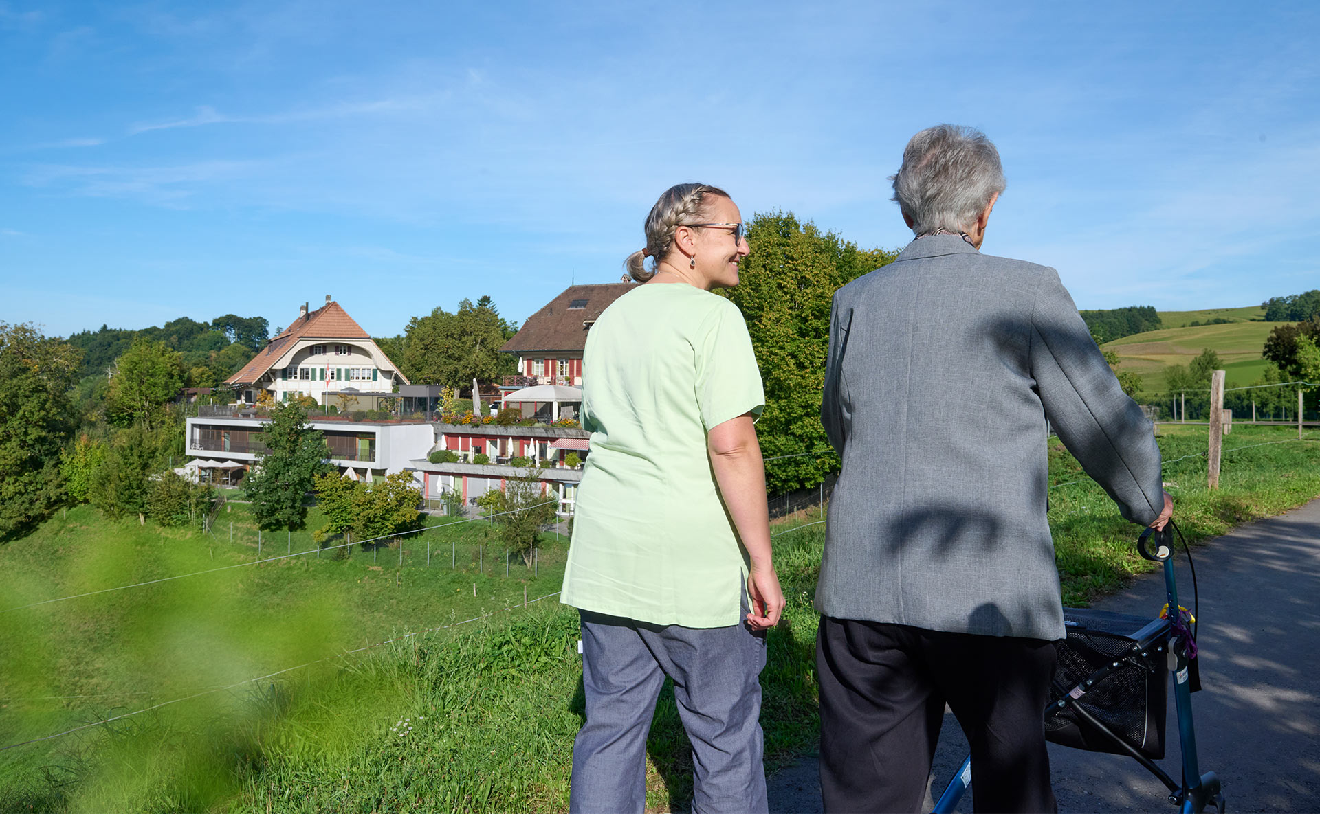Eine Bewohnerin mit Rollator spaziert mit einer Pflegerin, im Hintergrund die ländliche Umgebung des Landhaus Neuenegg